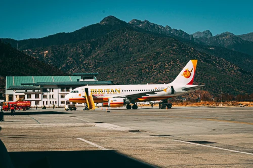 A Bhutan Airways Airbus A319 is parked at the apron with mountainous scenery in the background.