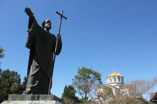 A bronze statue of a saint holding a cross, with a church dome under a clear blue sky