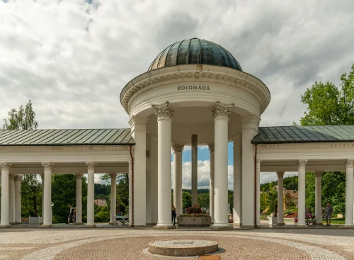 A scenic view of an elegant neo-classical colonnade structure with a domed roof, surrounded by lush greenery and blue sky