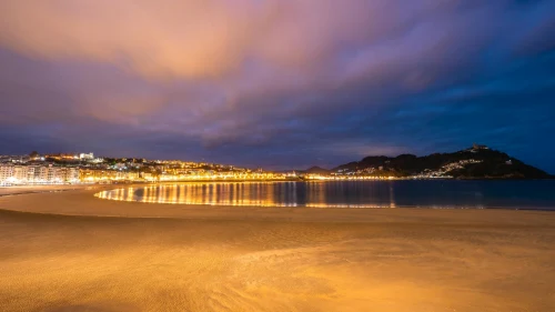 A stunning view of San Sebastian beach at twilight, highlighting city lights reflecting on the ocean under a dramatic sky