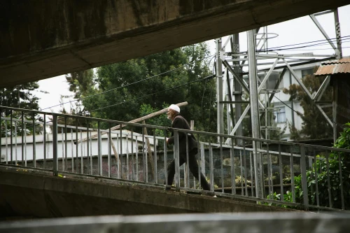 Elderly man with cane walks on an overpass in a bustling urban area, surrounded by greenery