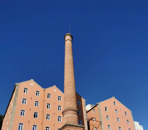 A tall brick smokestack with pink buildings against a clear blue sky in Porto, Portugal
