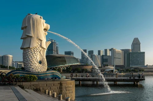 Iconic Merlion statue with Singapore skyline under clear blue sky, showcasing modern architecture and city's charm
