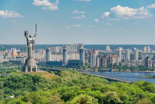 Motherland Monument surrounded with lush green trees on embankment of river near tall buildings under bright blue sky with clouds